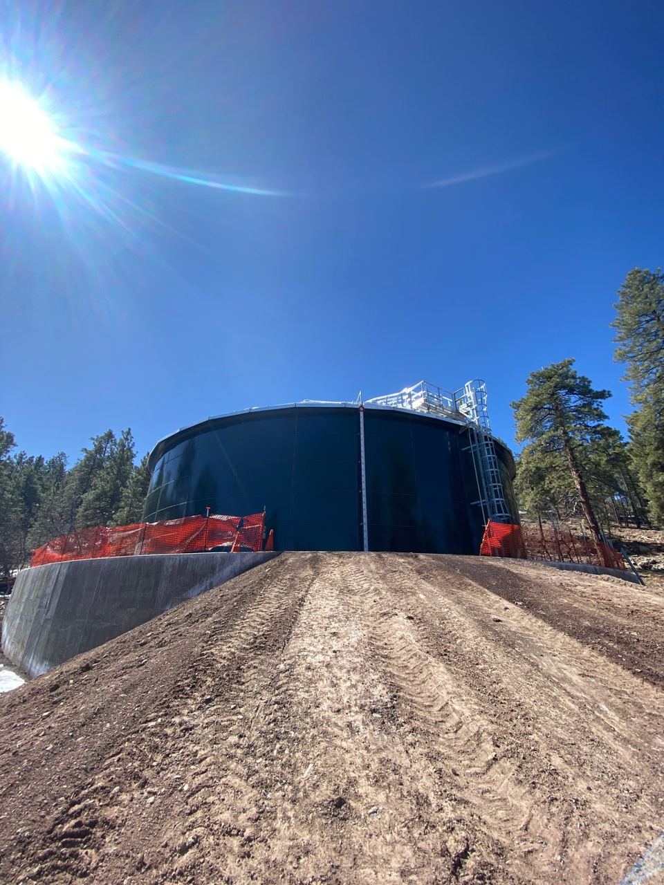 Dark blue bolted steel tank on an engineered earthen berm at a forested mountain site
