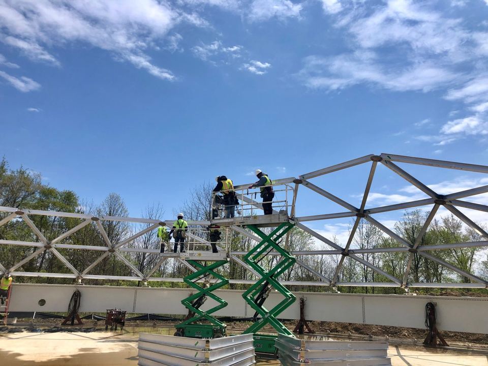 Crew on a scissor lift assembling aluminum dome roof rafters atop a white bolted steel tank wall
