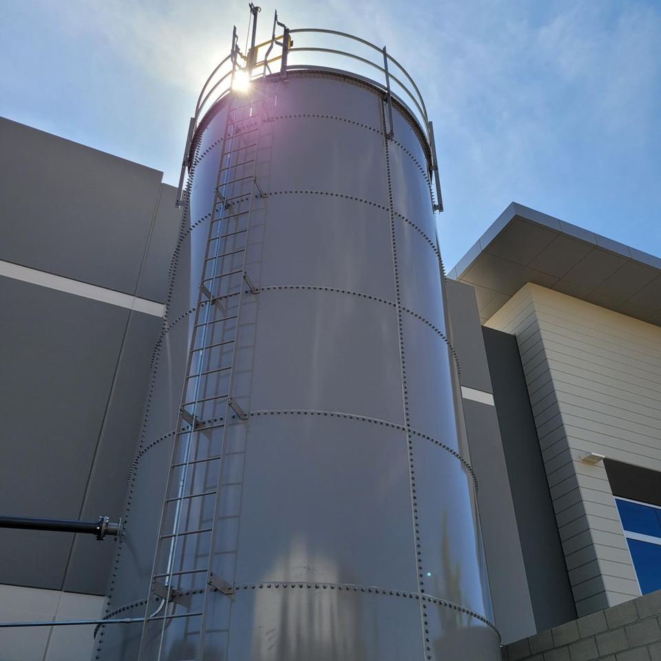 Tall grey bolted steel tank with caged exterior ladder beside an industrial building