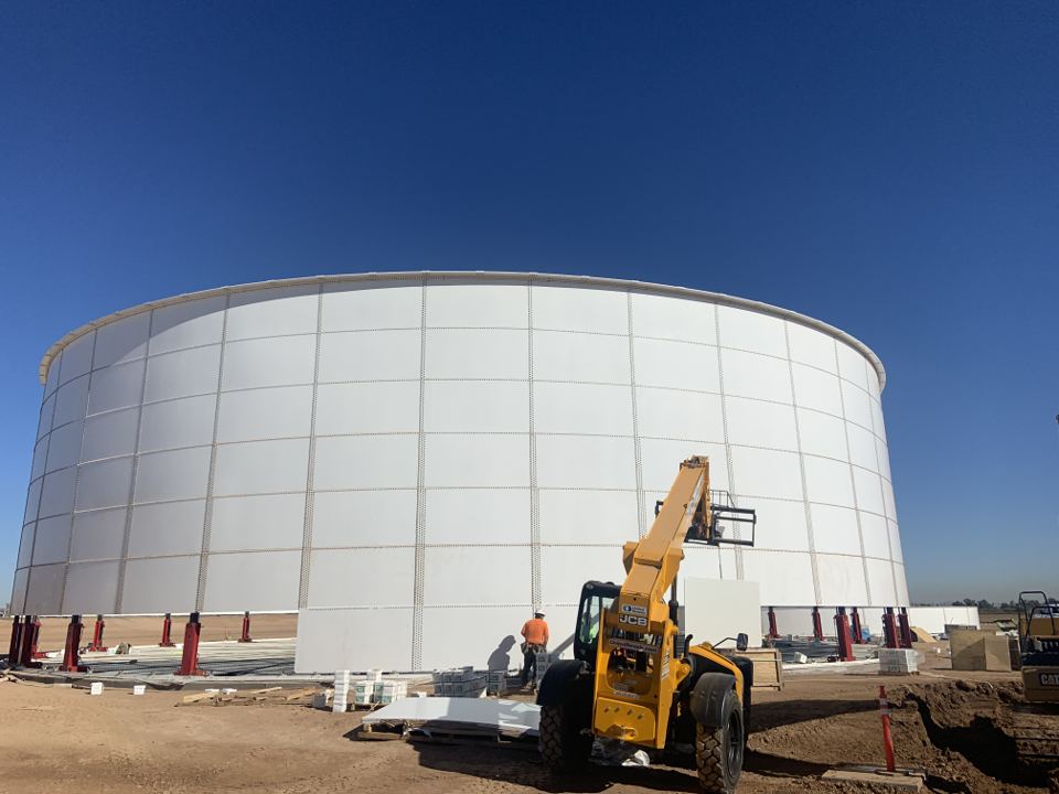 Large white bolted steel tank shell raised on hydraulic jacks during erection with a telehandler alongside