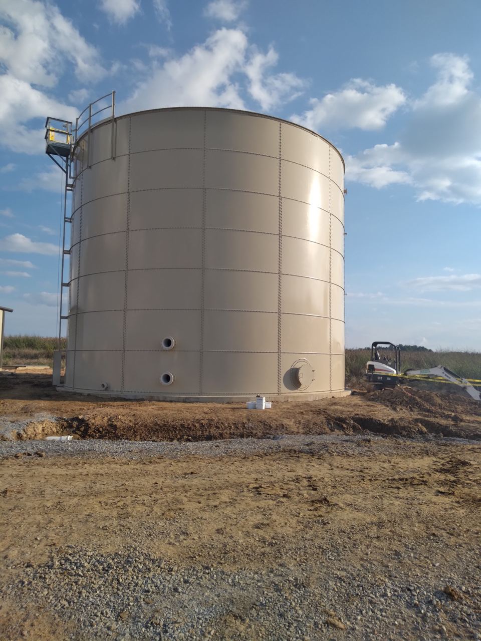 Completed tan bolted steel tank with exterior ladder and nozzle penetrations under a blue sky