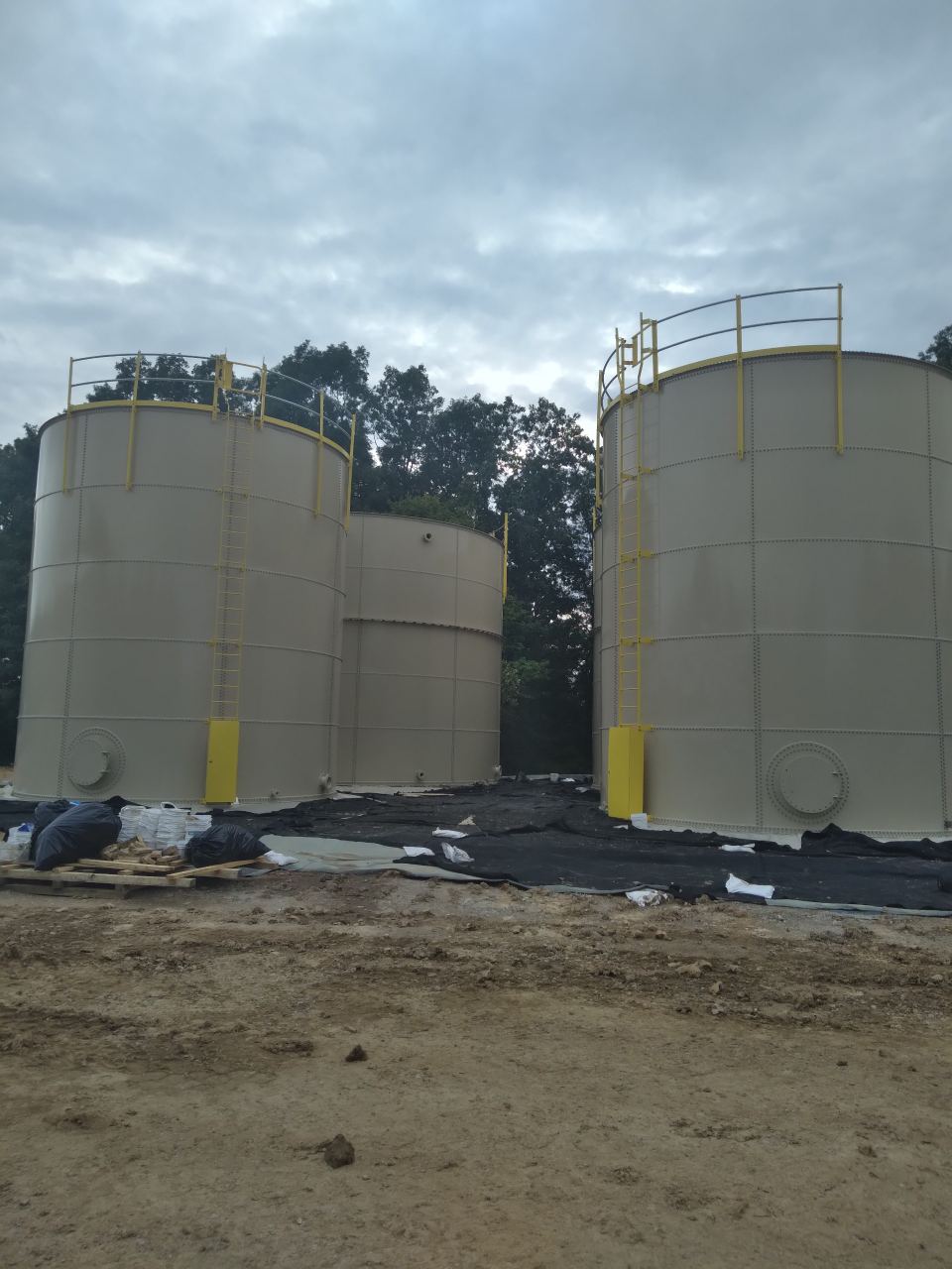 Pair of tan bolted steel tanks with safety handrails beside a geomembrane-lined containment area