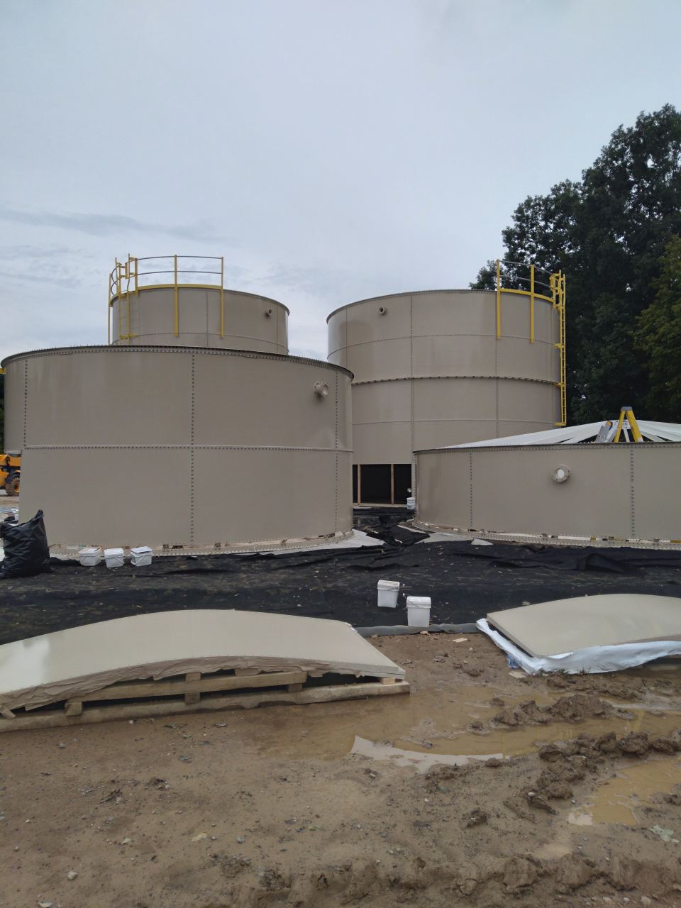 Three tan bolted steel tanks under construction with roof panels staged on the ground