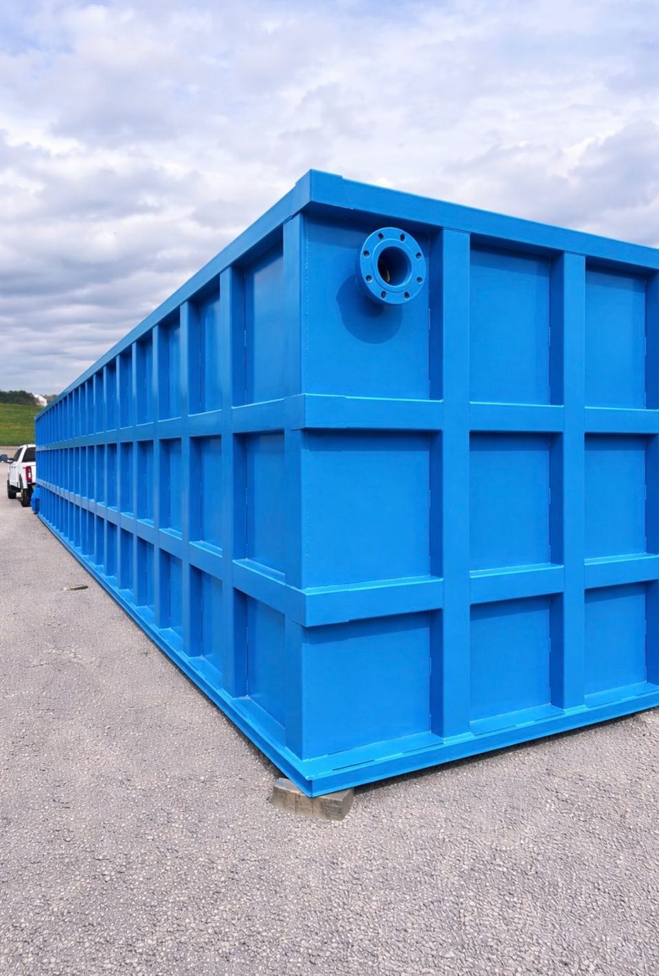 Long blue modular steel tank staged outdoors in the yard, top-corner flanged nozzle and reinforced panel grid receding to the horizon