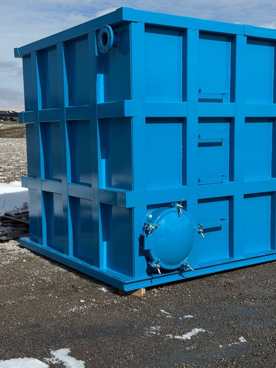 Corner of a blue modular steel tank with a bolted, gasketed manway and wing-nut clamps, staged on a winter lot