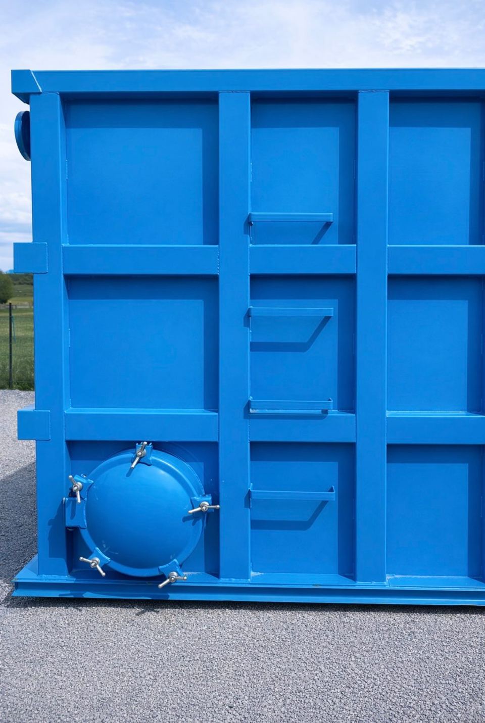 End wall of a blue modular steel tank with a bolted circular manway and welded step brackets, outdoors on gravel