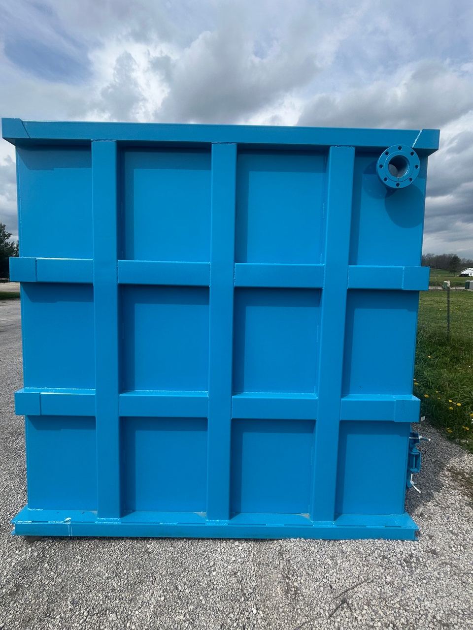 End wall of a finished blue modular steel tank with its reinforced panel grid and a corner flanged nozzle, outdoors under a cloudy sky