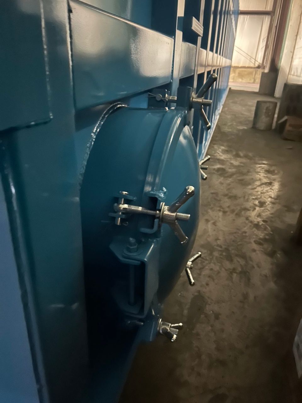 Close-up of a side-mounted bolted manway with swing-bolt wing-nut clamps on a blue modular steel tank in the shop