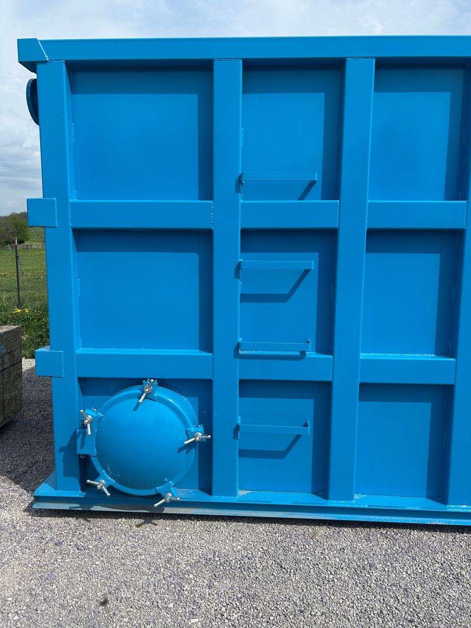 End wall of a blue modular steel tank with a bolted manway and welded step brackets for access, staged outdoors