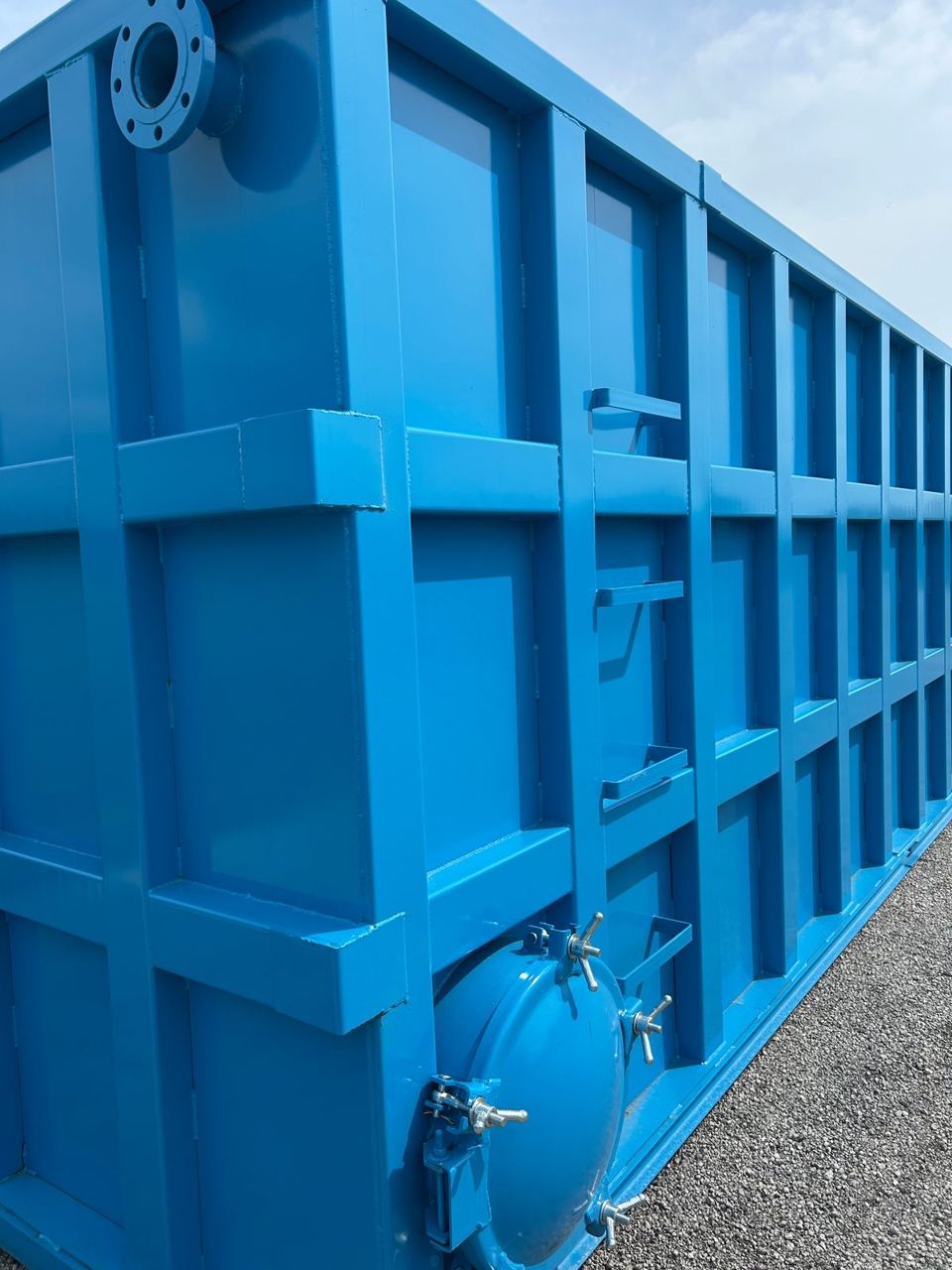 Long side of a blue modular steel tank outdoors showing the reinforced panel grid, top nozzle, and a side-mounted bolted manway