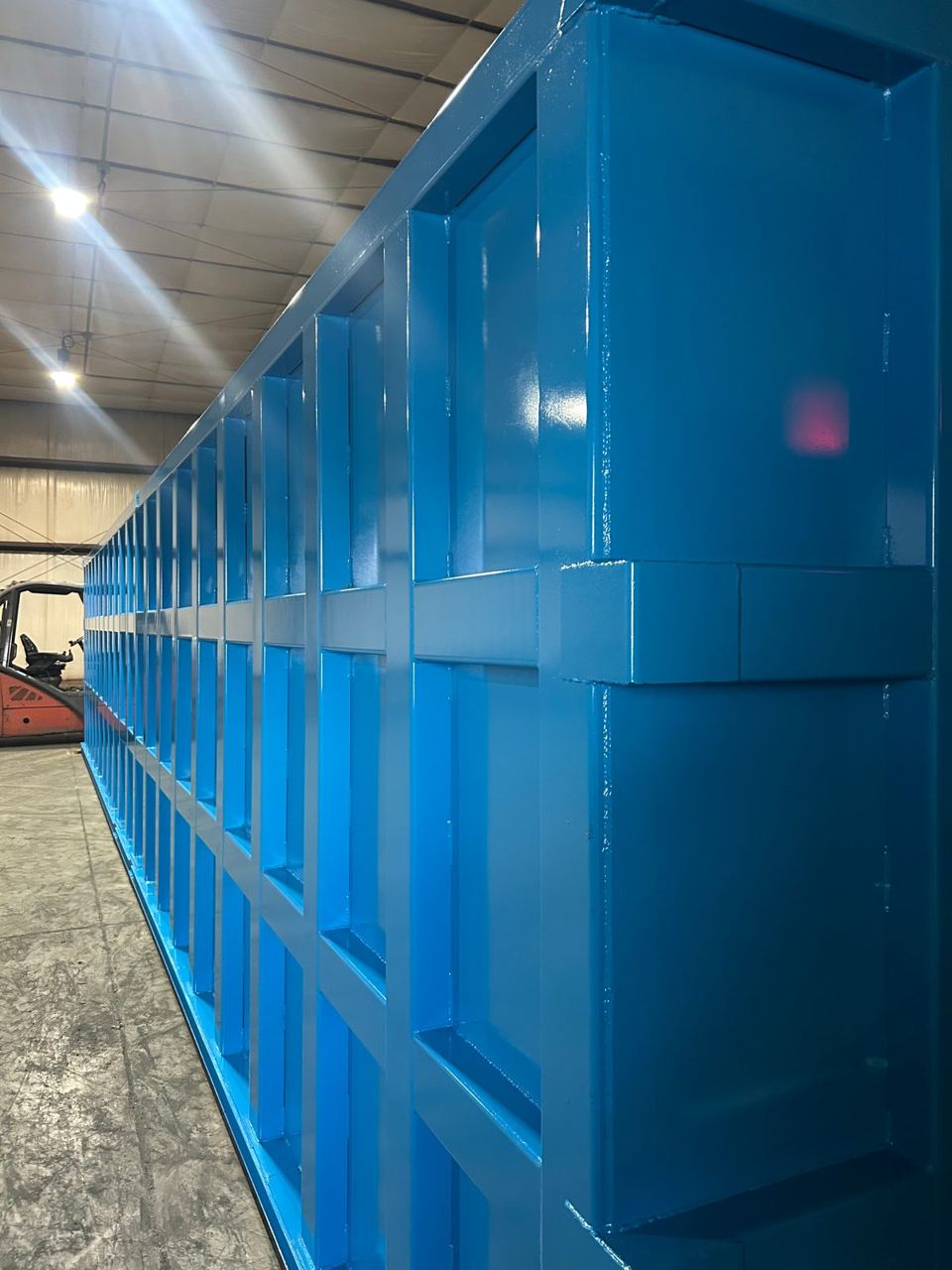 Freshly coated blue modular steel tank running the length of the fabrication shop, glossy panels reflecting the overhead lights