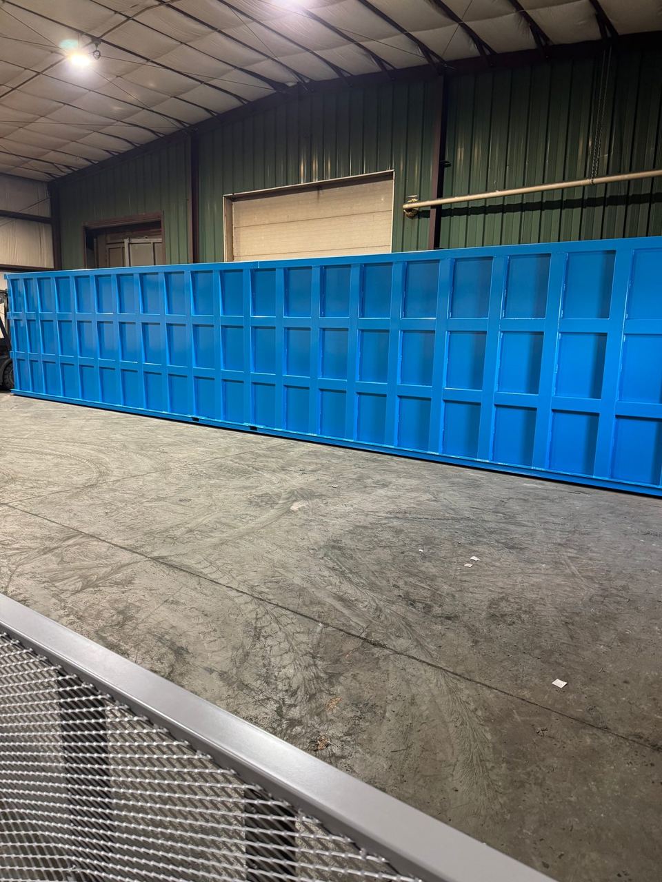 Long low blue modular steel tank in the shop, its full reinforced panel wall facing the camera