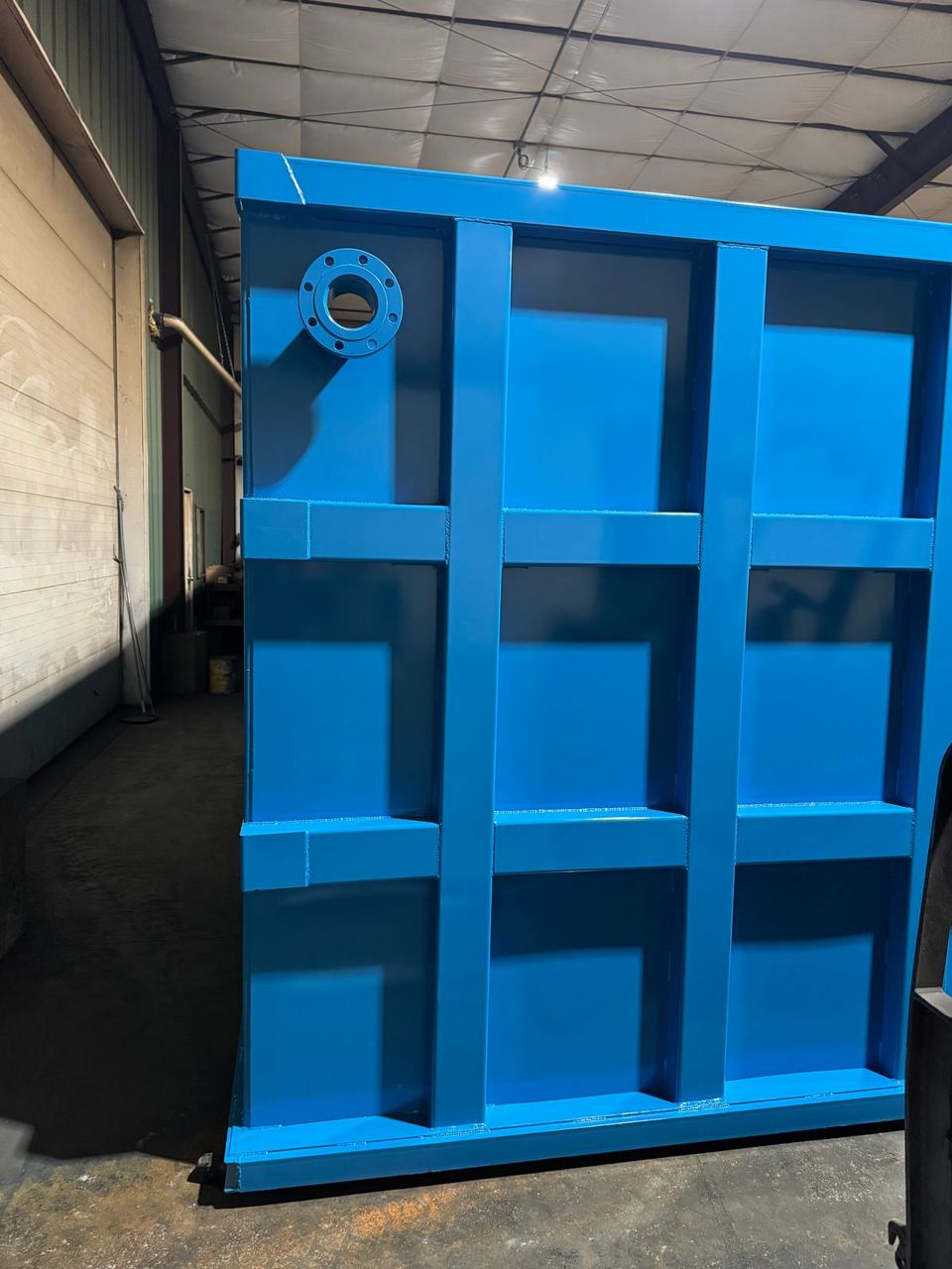 Tall blue modular steel tank end section with a flanged nozzle and three-by-three reinforced panel grid inside the fabrication shop