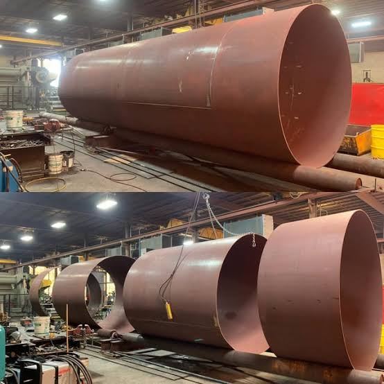 Primer-coated carbon steel round tank lying in the fabrication shop with its open end toward the camera, above a row of rolled shell courses staged for seam welding
