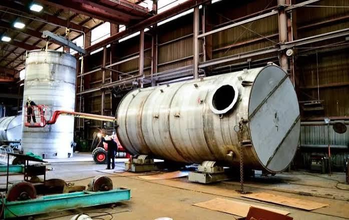 Stainless steel shop-welded round tanks under fabrication &mdash; a large horizontal tank on turning rolls with an open manway nozzle, and a vertical tank being lifted by overhead crane behind it