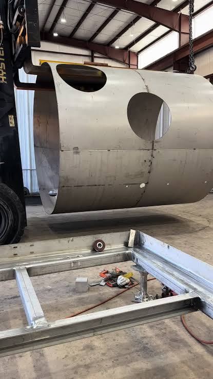Stainless steel round tank shell section with circular nozzle cutouts, held on a forklift boom above its steel base frame on the shop floor