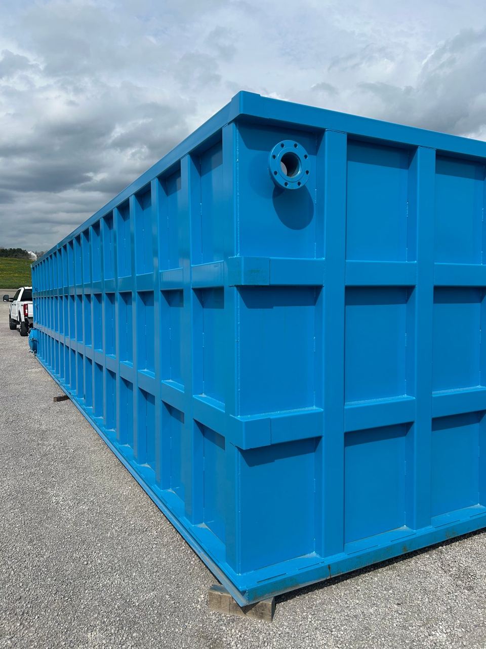 Large blue shop-welded steel tank staged outdoors with a pickup truck alongside for scale, top nozzle and bolted panel seams