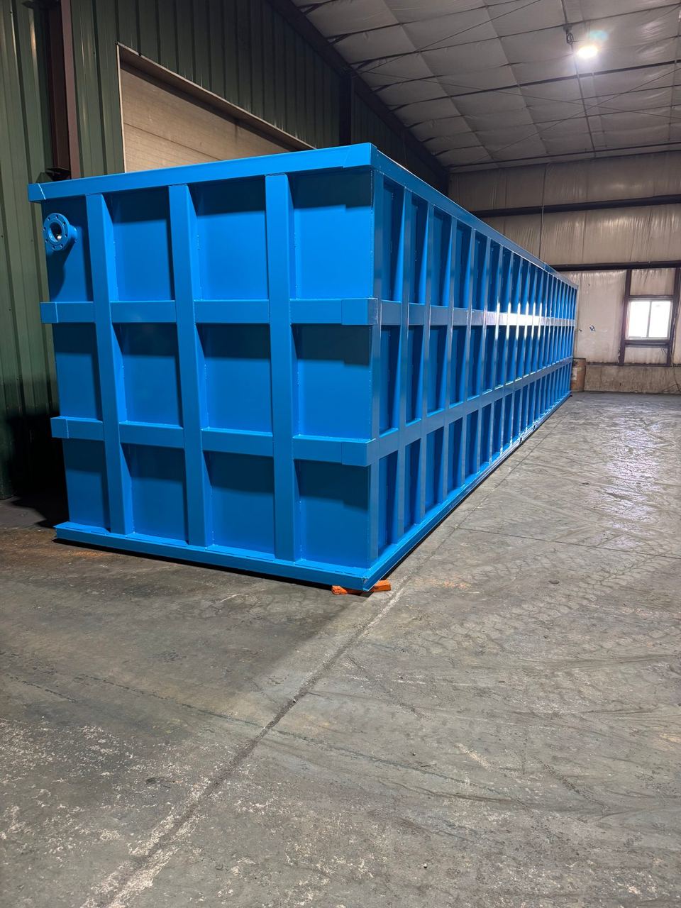 Three-quarter view of a long blue shop-welded steel tank in the shop with a welded lifting lug at the corner