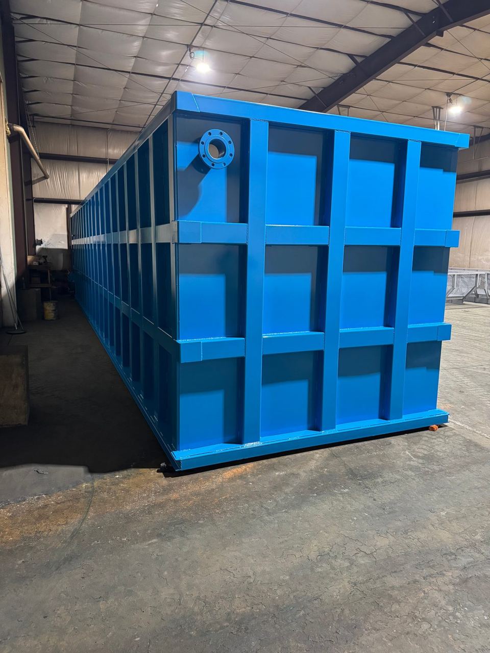 Long blue shop-welded steel tank in the shop with a top-corner flanged nozzle and the reinforced panel grid running its full length