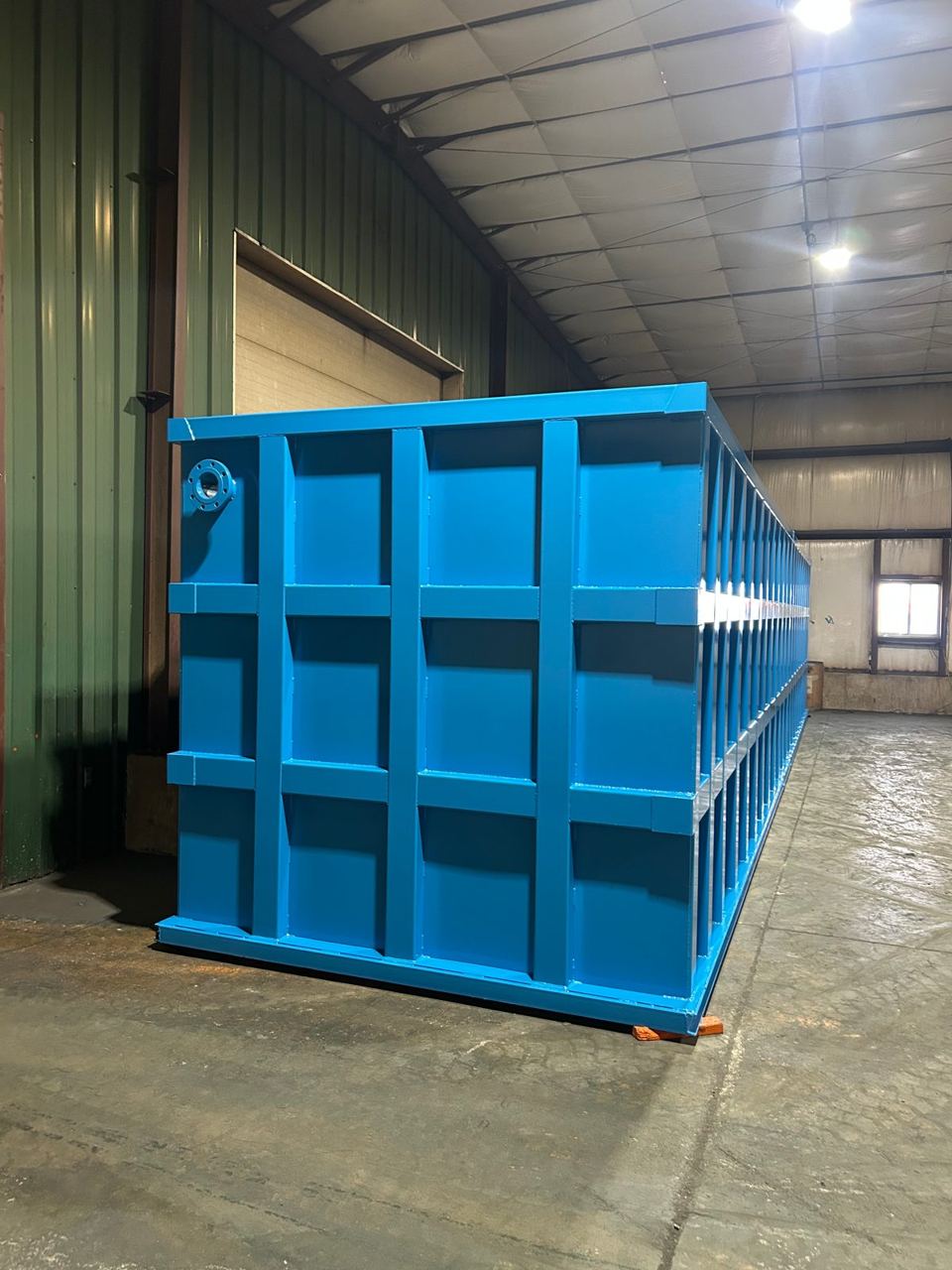 Three-quarter view of a long blue shop-welded steel tank inside the shop, lifting lug on the near corner