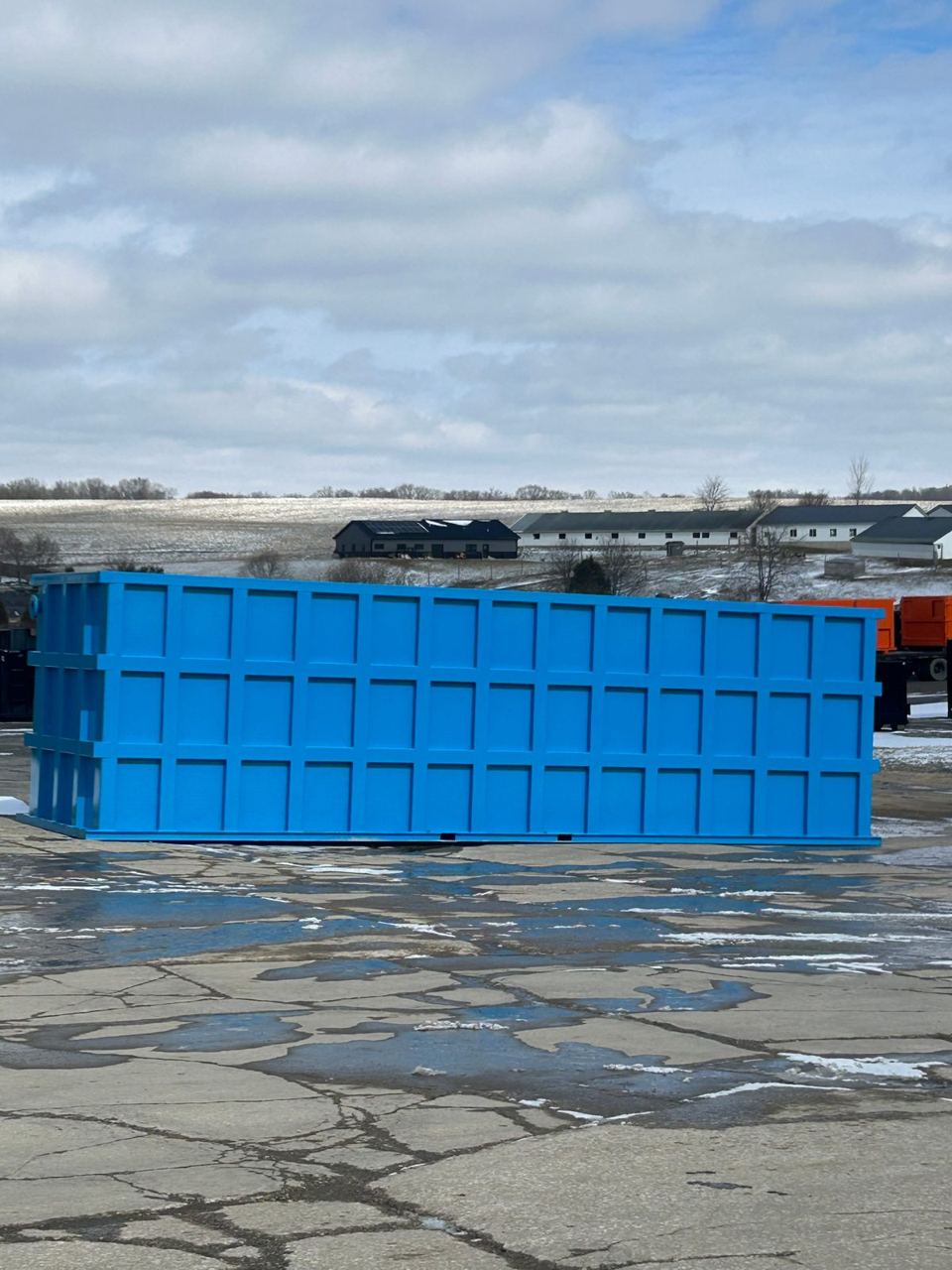Blue shop-welded steel tank staged on a snow-dusted yard with farm buildings on the horizon