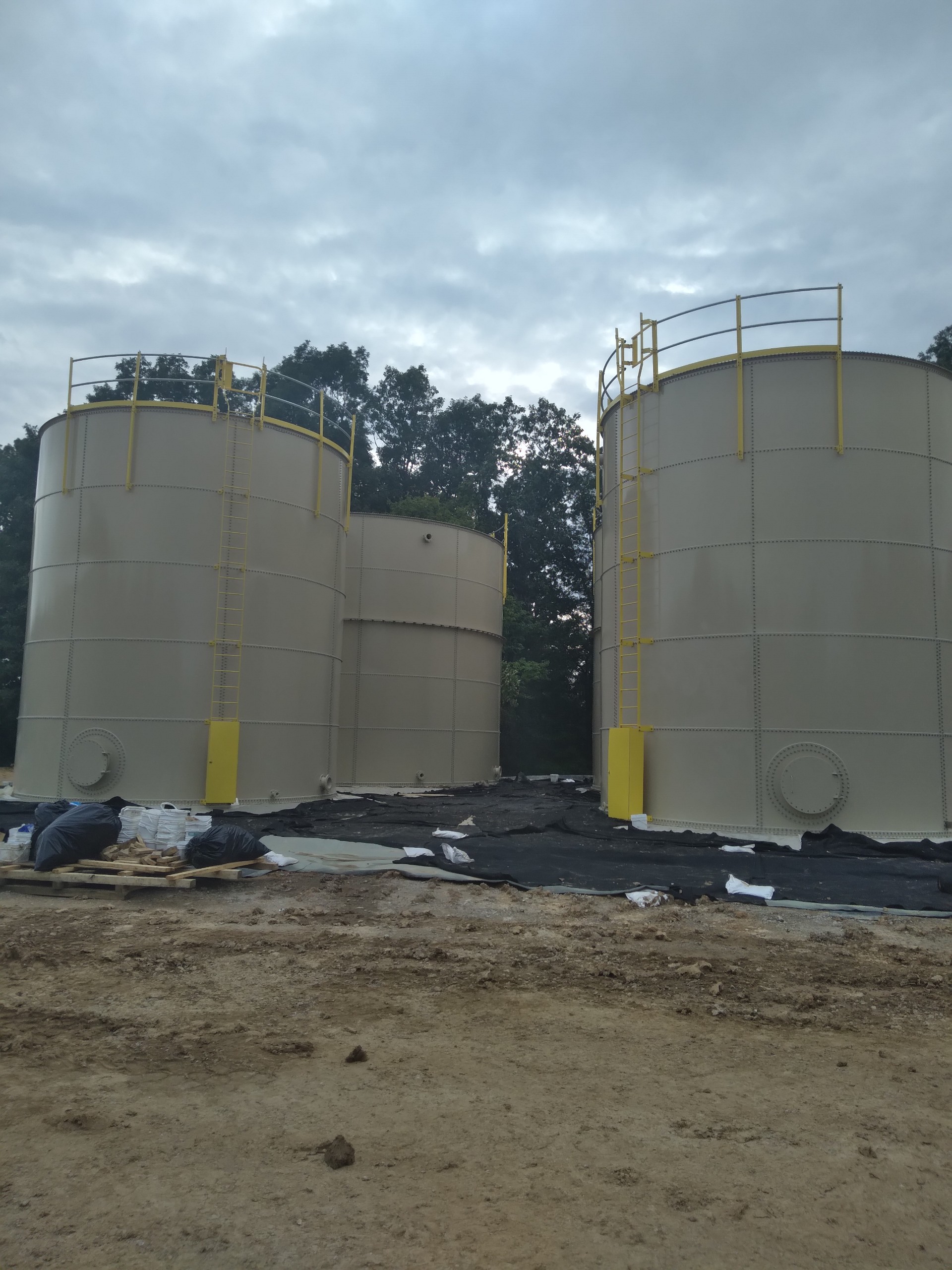 Pair of tan bolted steel tanks with safety handrails beside a geomembrane-lined containment area
