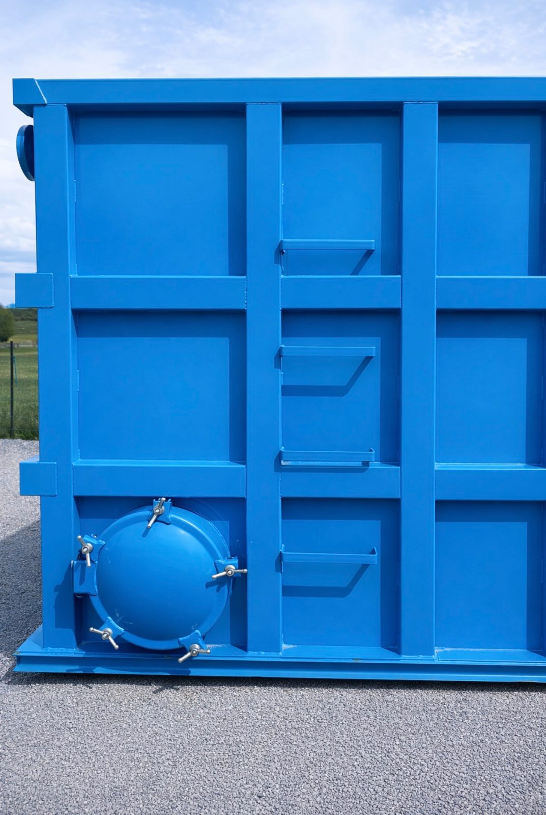 End wall of a blue modular steel tank with a bolted circular manway and welded step brackets, outdoors on gravel