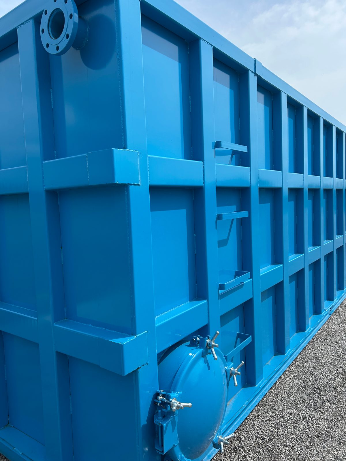 Long side of a blue modular steel tank outdoors showing the reinforced panel grid, top nozzle, and a side-mounted bolted manway