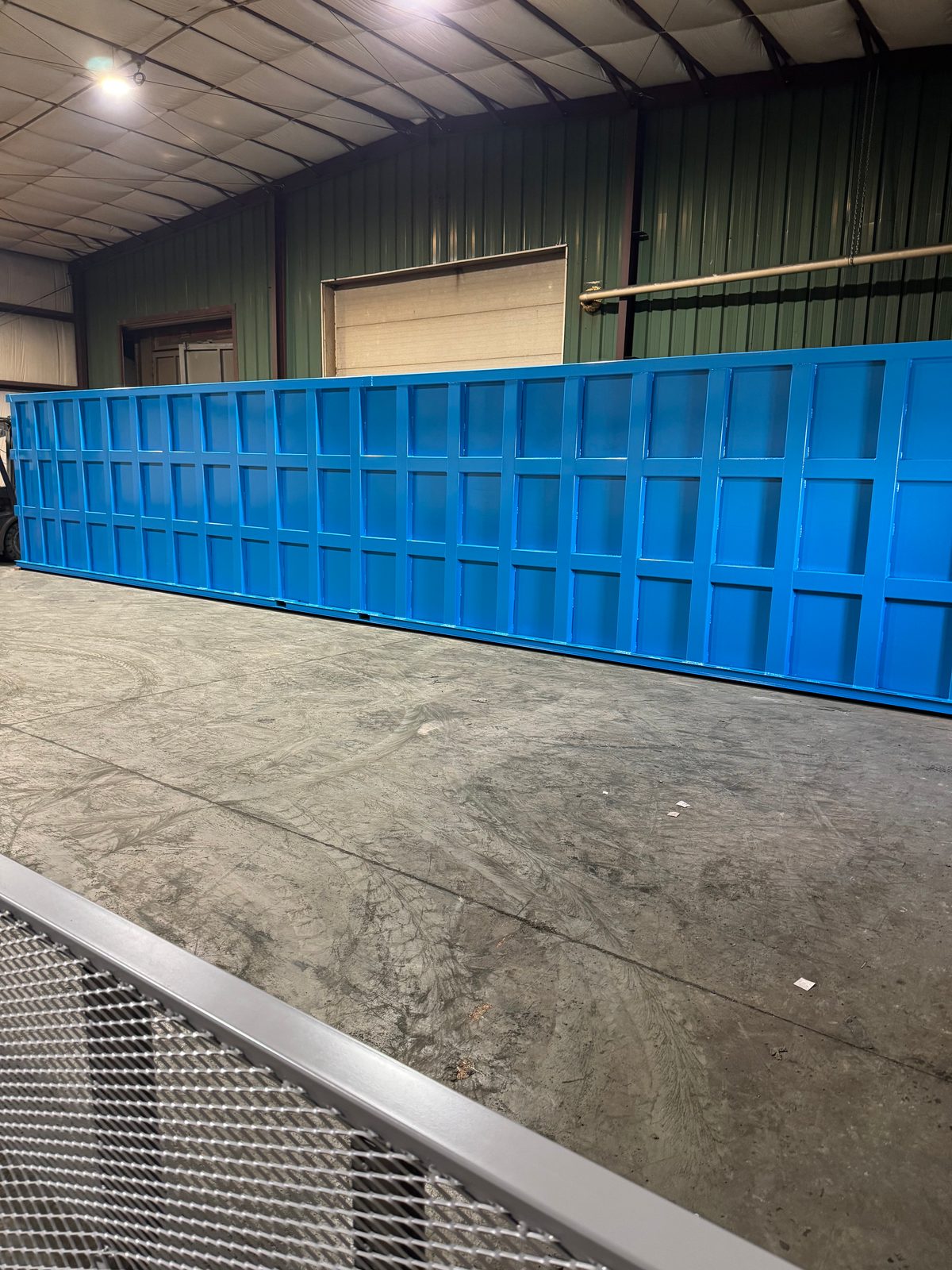 Long low blue modular steel tank in the shop, its full reinforced panel wall facing the camera